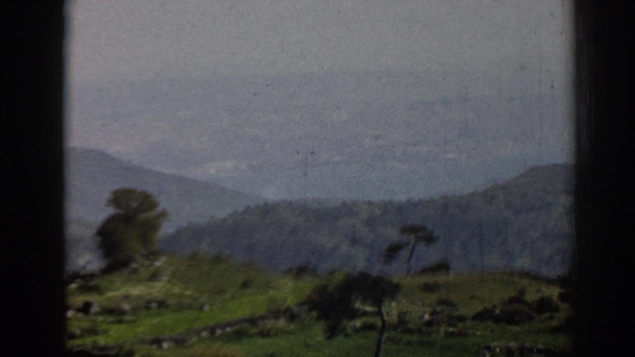 1962 home-movie frame near Ephesus showing a closer hillside view with trees, walls, and soft mountain background.