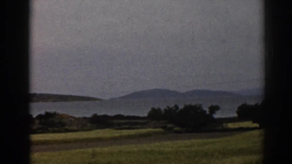 1962 8mm view near Ephesus, Turkey, with fields, shrubs, water, and distant hills passing behind dark frame or window edges.