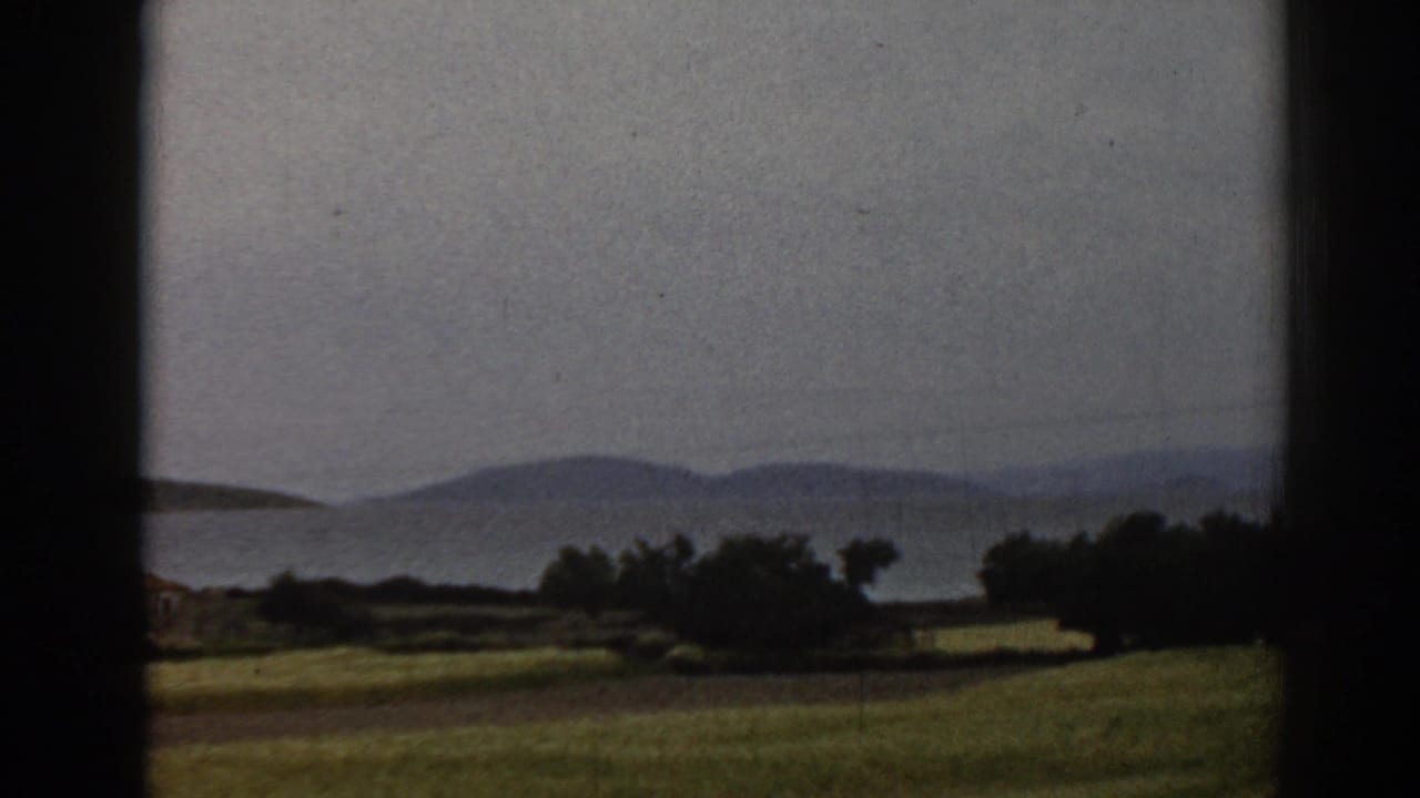 1962 8mm travel footage near Ephesus, Turkey, with a grassy slope, shoreline vegetation, water, and layered hills in soft focus.