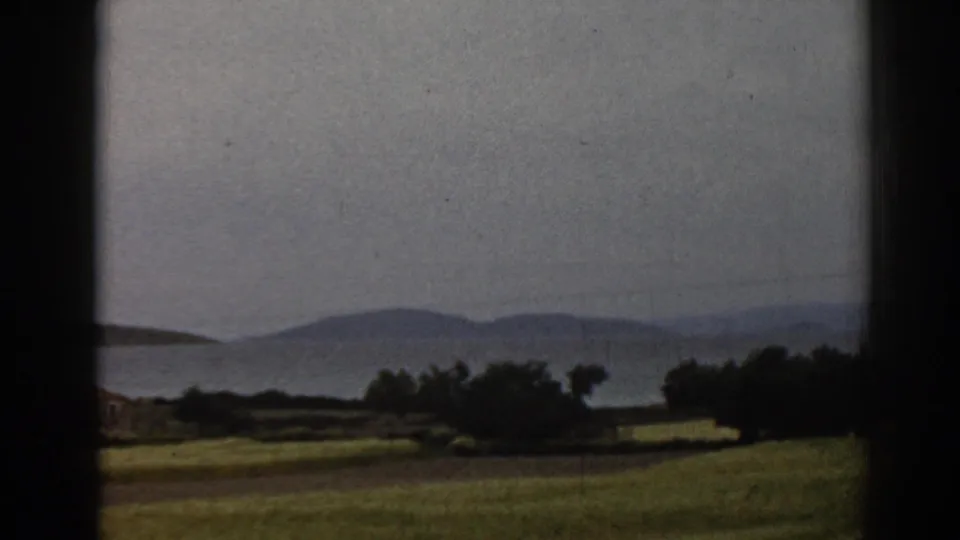 1962 8mm travel footage near Ephesus, Turkey, with a grassy slope, shoreline vegetation, water, and layered hills in soft focus.