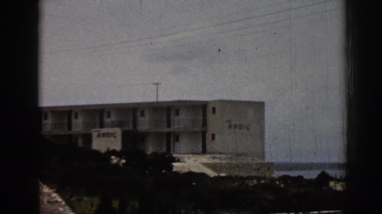 1962 Turkey home-movie frame showing a distant cliffside hotel with balconies, shrubs, and sea in the background.