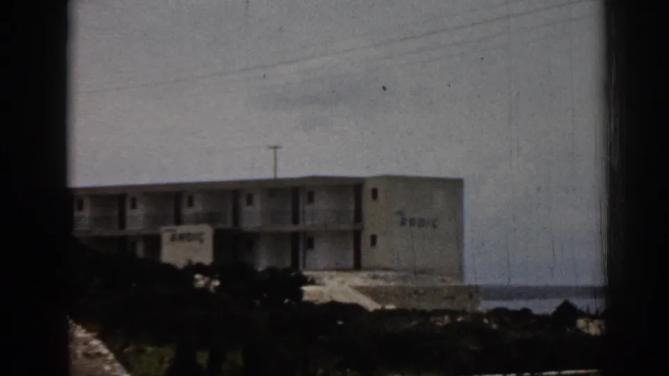 1962 Turkey home-movie frame showing a distant cliffside hotel with balconies, shrubs, and sea in the background.