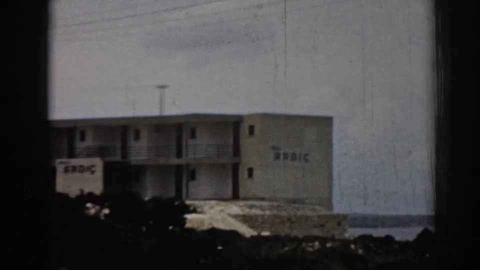 Closer 1962 Turkey frame of the ARDIC-marked hotel, with balcony railings, rocky foreground, and water beyond.