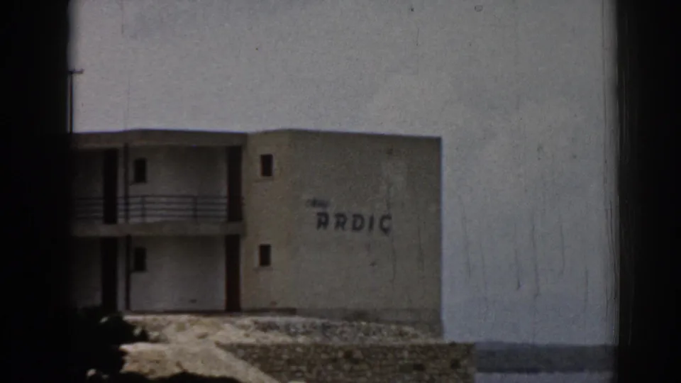 Tight 1962 Turkey home-movie frame of the ARDIC-marked hotel wall, balconies, rocky foreground, and distant water.