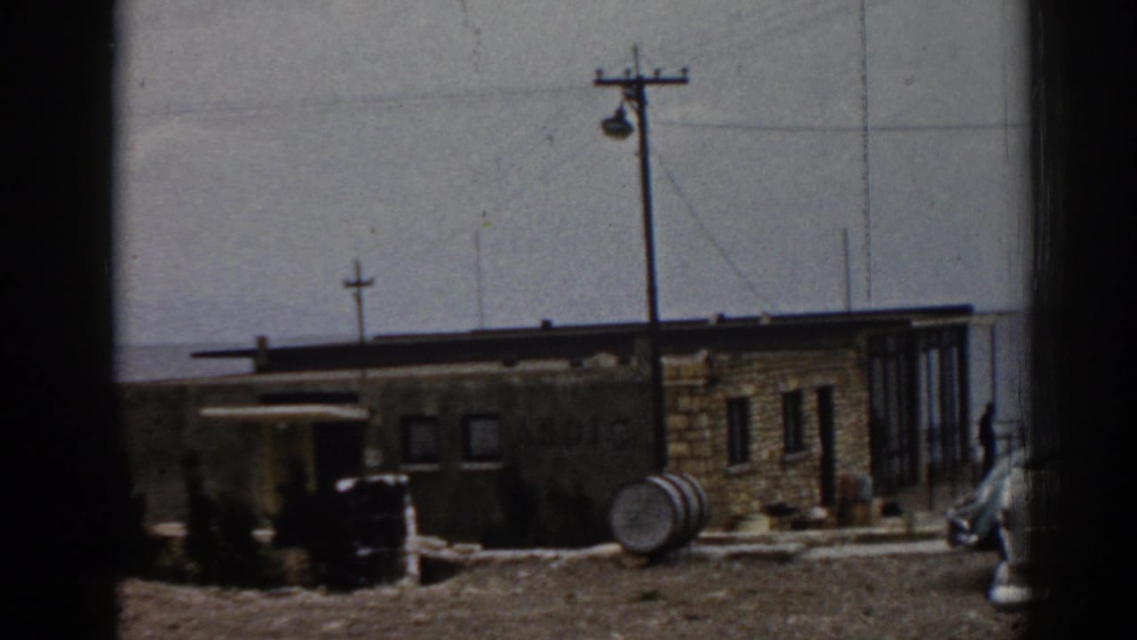 Tan stone building with a utility pole, barrel, and parked vehicle edge under a clear sky.