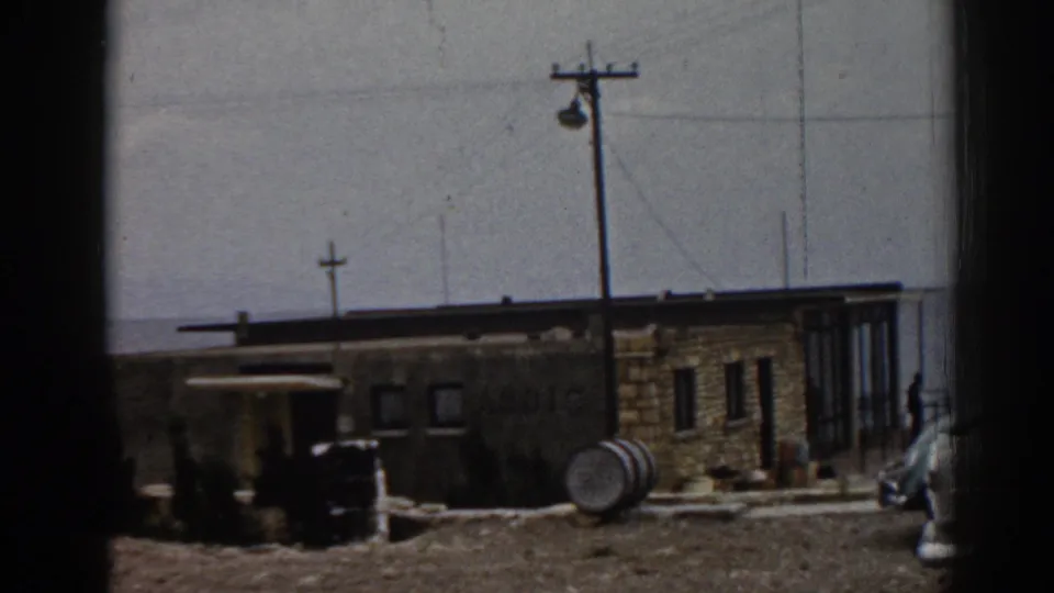 1962 Ephesus home-movie frame showing a tan stone building, utility pole, barrel, open ground, and partial parked car.