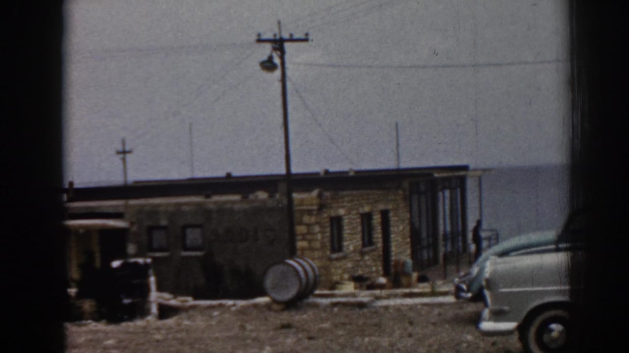 White car parked beside a tan building with glass windows, barrel, utility pole, and distant figure.