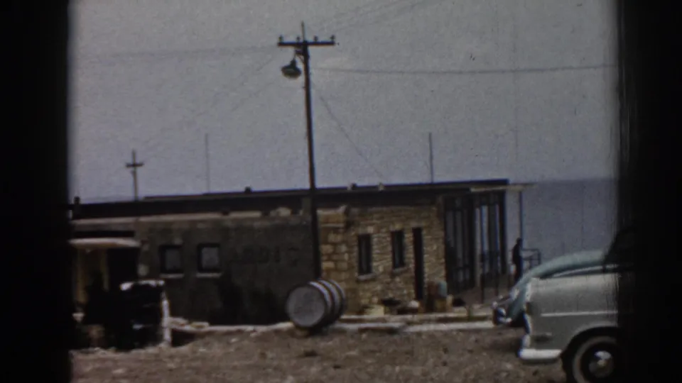 1962 Ephesus frame with a white classic car parked near a tan stone building, glass panels, utility pole, barrel, and small standing figure.