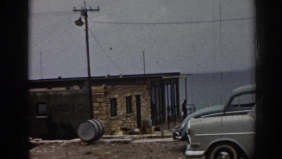 1962 Ephesus home-movie view of a white classic car parked beside a low tan building with a barrel and utility pole in front.