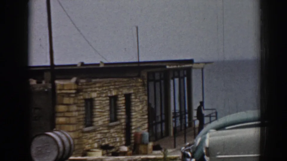 1962 Ephesus frame with a closer view of the tan stone building, white car, glass entrance, barrel, and distant person near a railing.