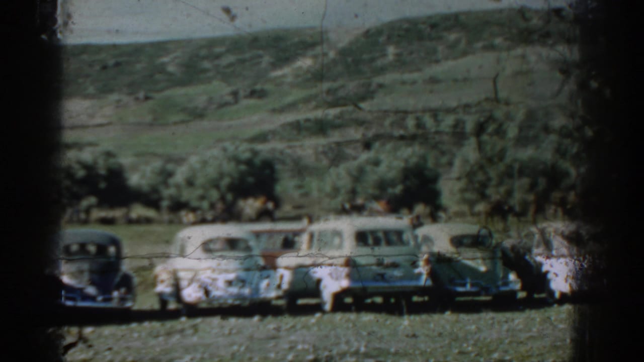 Parked vintage cars in a grassy rural field with trees and hills behind them.