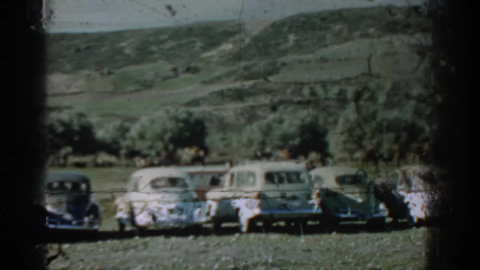 1962 Izmir home-movie frame showing a row of vintage cars parked in a grassy field below scattered trees and rolling hills.