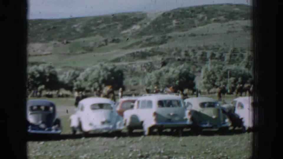 1962 Izmir rural scene with classic cars lined up on grass, distant people near the vehicles, trees, and rolling hills behind them.