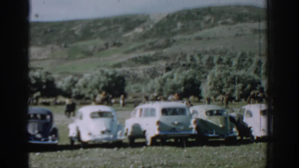 1962 Izmir home-movie frame of parked vintage cars in a grassy rural field, with small people visible near the cars and hills beyond.