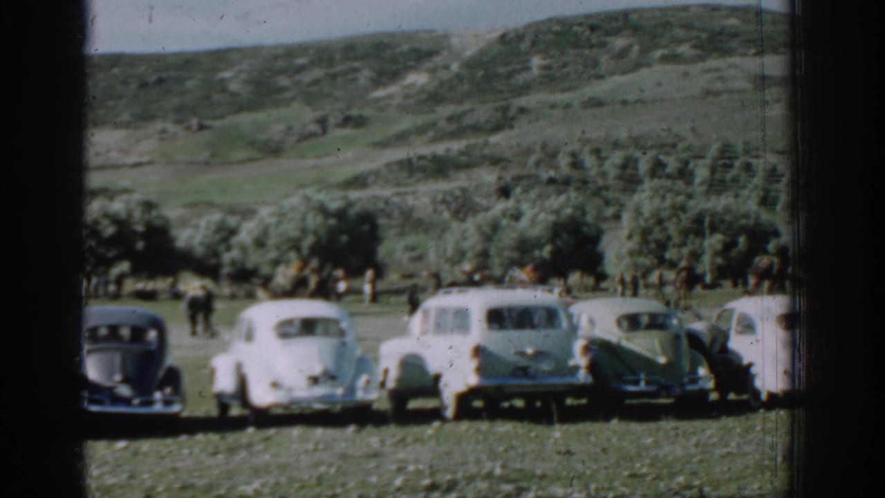 1962 Izmir home-movie frame showing several rounded cars parked in a grassy field beneath low hills and trees.