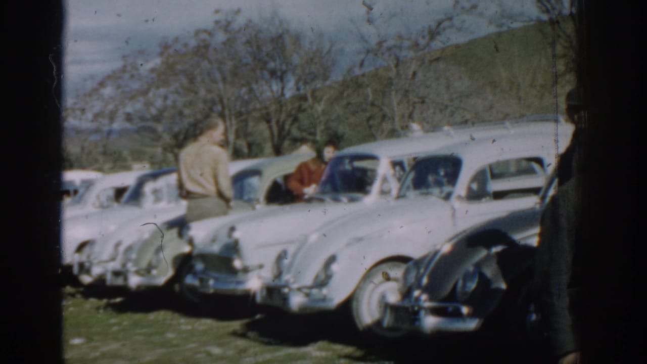 Adults stand beside a row of parked cars in a hilly field.
