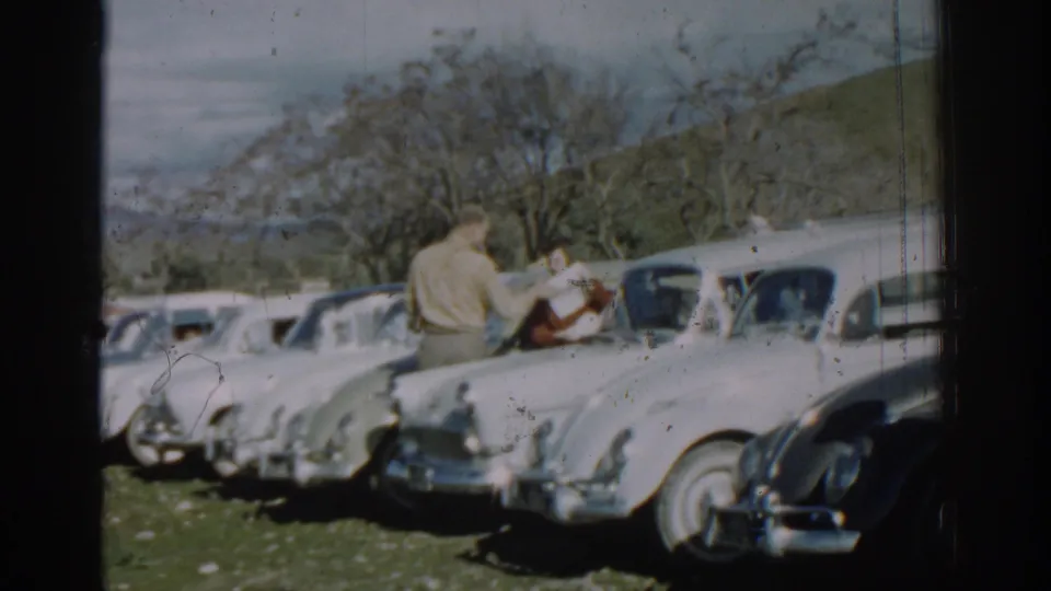 1962 Izmir home-movie frame showing a khaki-shirted man and a woman in red near a line of parked cars in a field.