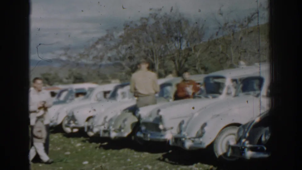 1962 Izmir frame with several adults around light-colored cars parked on grass, with trees and hills behind them.