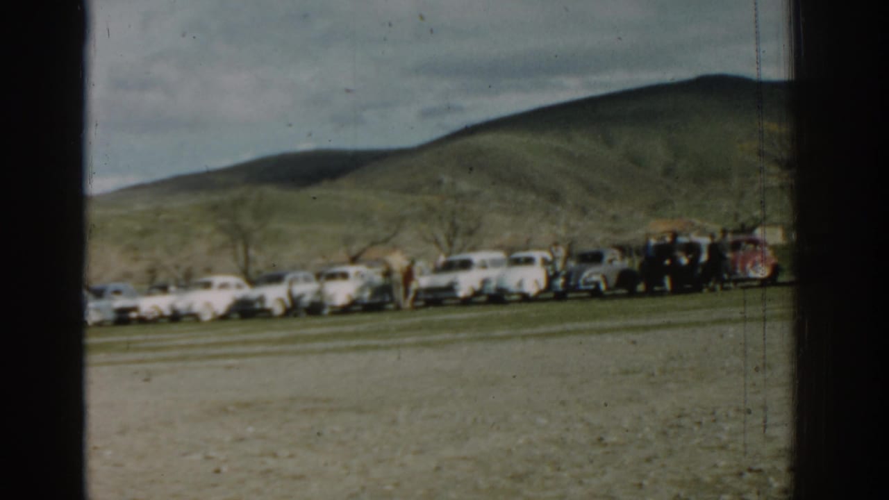 Row of light-colored cars parked near a hillside with a few people standing nearby.