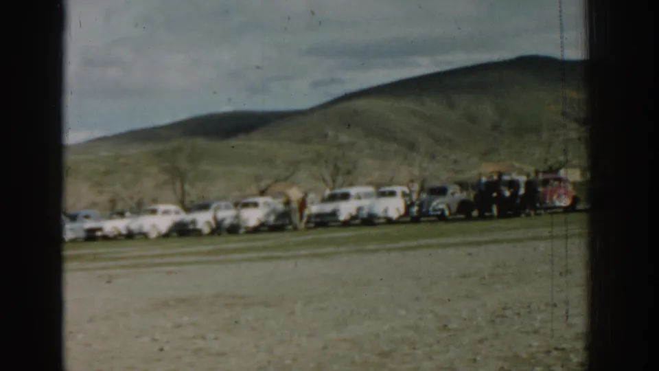 1962 8mm home-movie frame from Izmir showing pale cars parked in a row below rolling hills with distant bystanders.