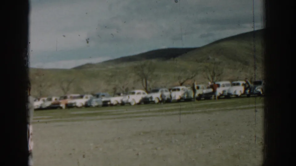 Soft 8mm frame showing parked mid-century cars across an open field in Izmir with small distant figures and hills.