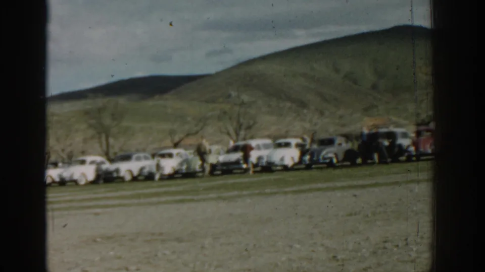 1962 Izmir home-movie frame with parked cars and distant people at the edge of a field below rolling hills.