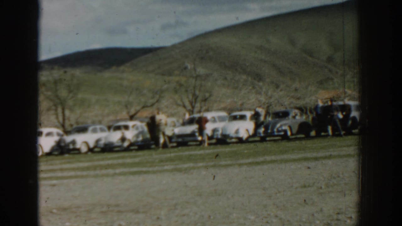 1962 Izmir home-movie frame showing classic cars parked in a line as adults gather near a grassy hillside.