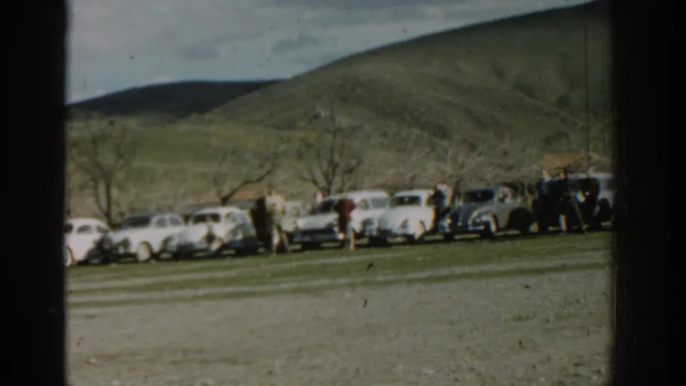 1962 Izmir home-movie frame showing classic cars parked in a line as adults gather near a grassy hillside.