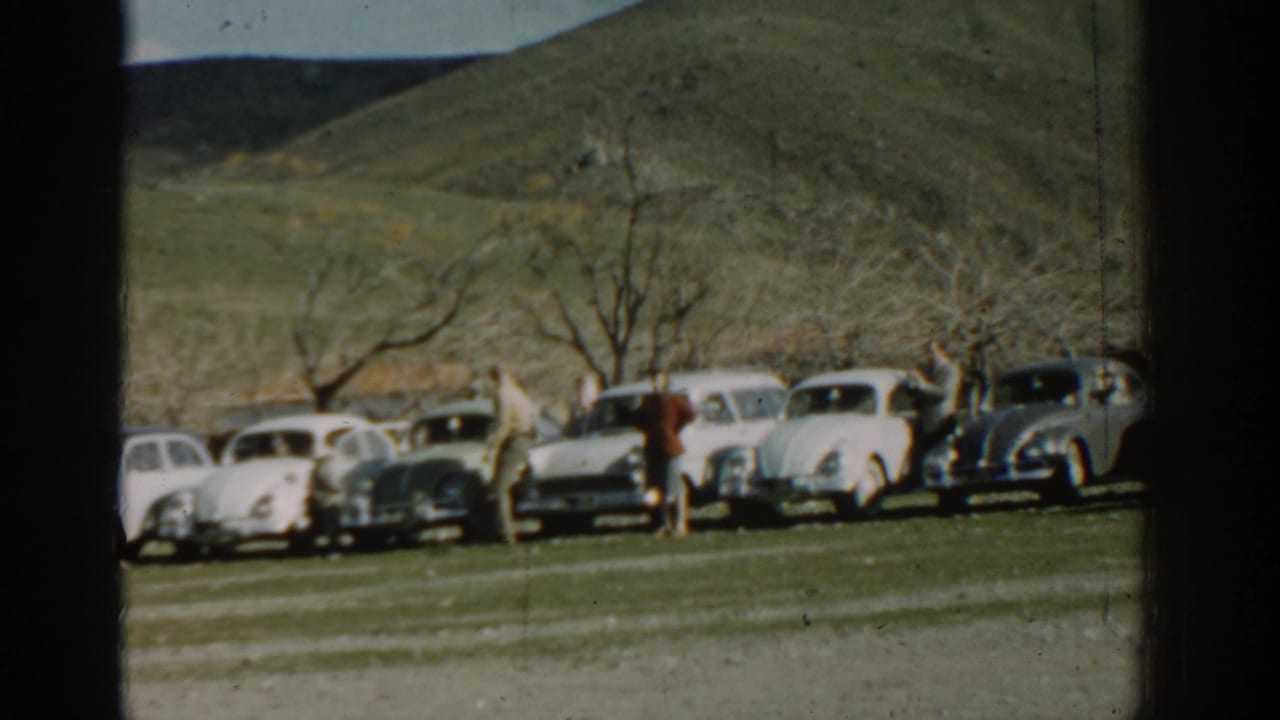 People walk beside parked vintage cars on grass with trees and hills in the background.