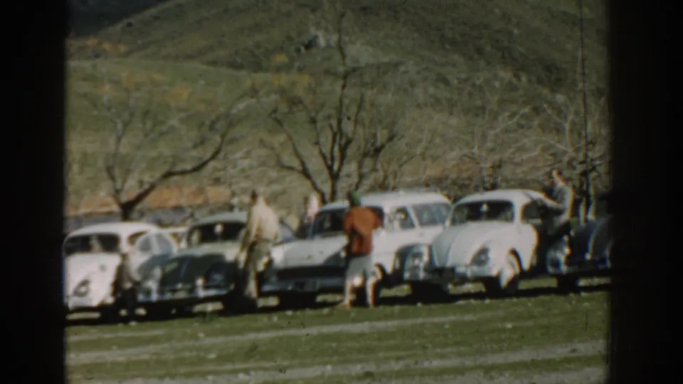 Adults in period clothing walk past several parked classic cars in this 1962 Izmir outdoor home-movie frame.