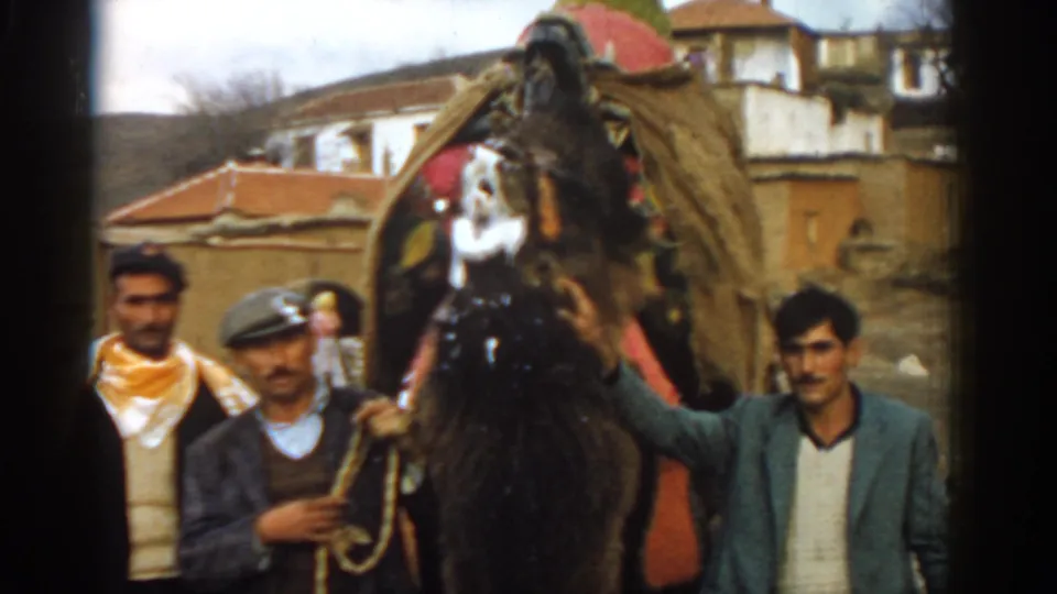 1962 Izmir-area home-movie frame with adult men standing beside the front of a decorated camel on an outdoor village road.