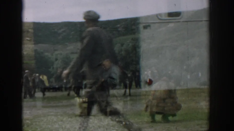 1962 Izmir frame with a blurred man crossing in front of a reflective view of men and large animals near a white vehicle or trailer.