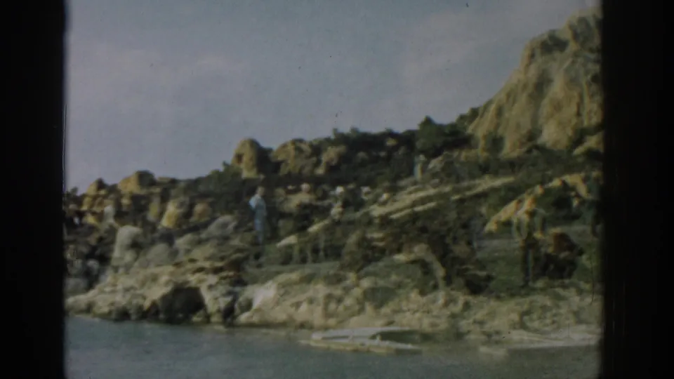 Wide 1962 Izmir home-movie frame of people on rocks watching a large unidentified animal by the water.