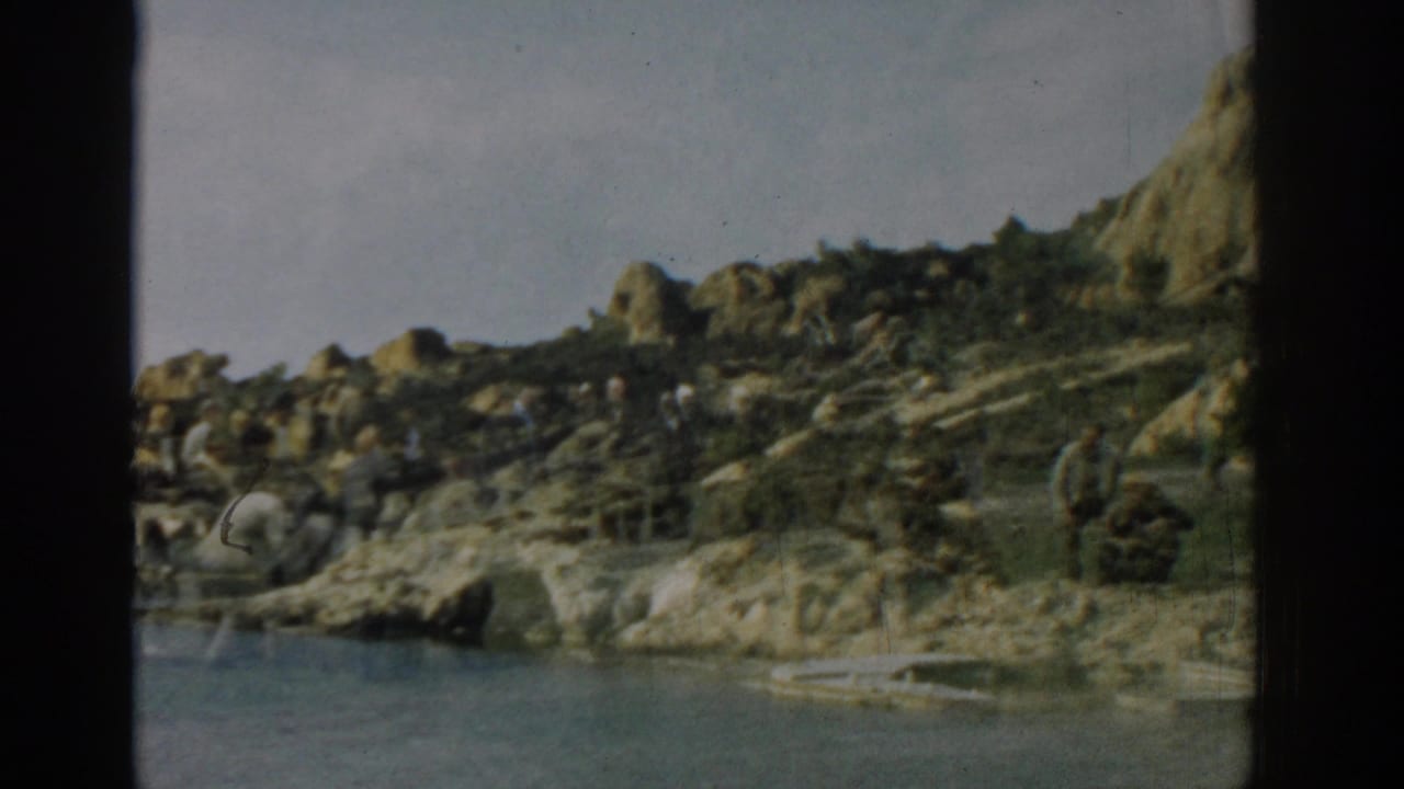 Rocky shoreline beside blue water with a small distant person near the rocks.