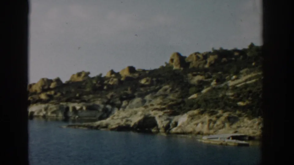 1962 Izmir coastal view with boulder-strewn rocks, calm sea, pale sky, and a small boat pulled close to the shoreline.