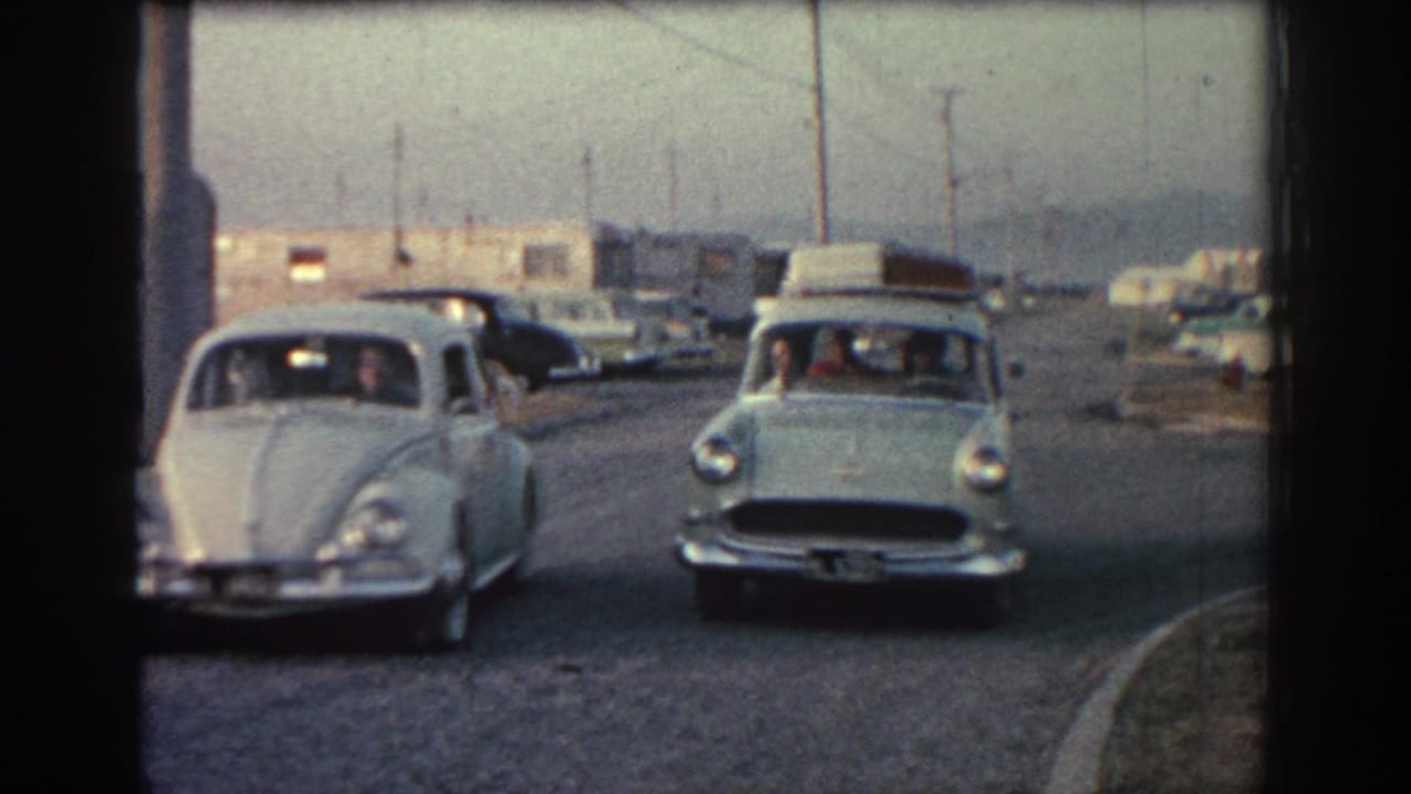 Two vintage cars approach along a road near trailers and utility poles.