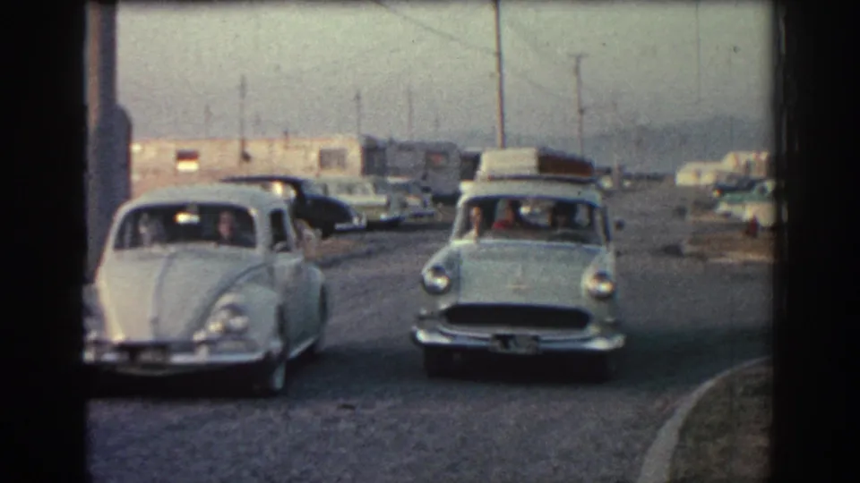 1962 Rhodes home-movie frame showing a white Volkswagen Beetle and another vintage car approaching a gravel roadside area.