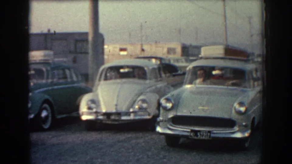 1962 Rhodes frame with a white Volkswagen Beetle and light green vintage car stopping beside other roadside vehicles.