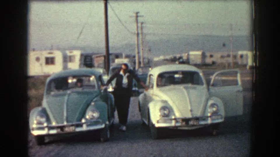 1962 Rhodes frame showing a man beside a green Beetle and white Beetle, with an open car door and trailers in the background.