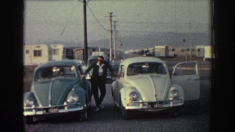 1962 Rhodes home-movie frame with a man standing near green and white Volkswagen Beetles in a gravel lot under a hazy sky.