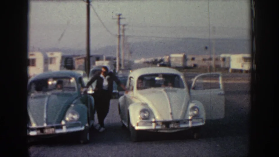 Rhodes, Greece home-movie frame of a man beside two parked cars at a roadside stop, with trailers and hazy hills in the distance.