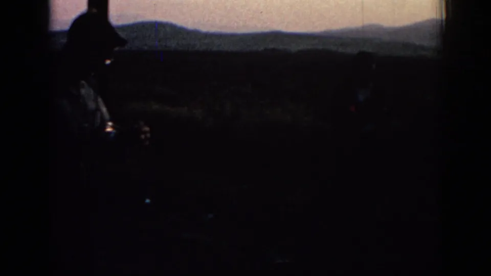 Dark 8mm frame showing a silhouetted roadside or countryside view with distant hills under a dim evening sky in the travel sequence.