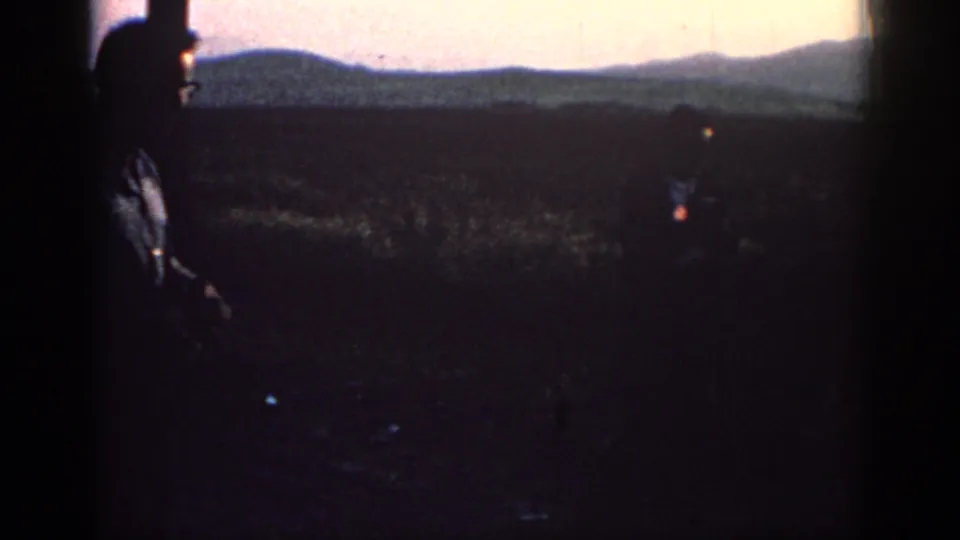 In Rhodes, Greece, 1962, two adult men appear silhouetted in a field at sunset, with distant hills on the horizon.