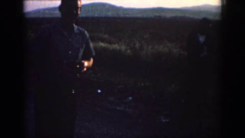 A silhouetted adult man stands in a Rhodes field at dusk, with another person partly cropped and hills behind them.