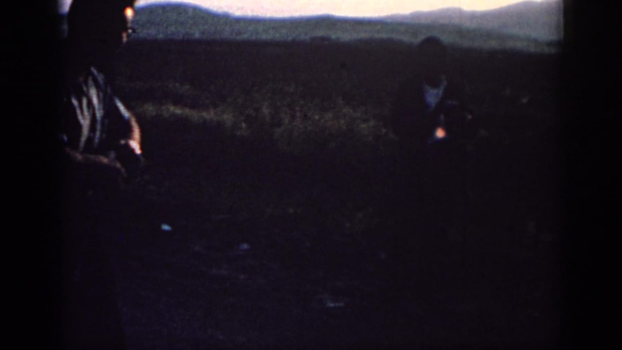 Two adult men appear in silhouette in a rural Rhodes field, with low hills and dim evening light in the background.