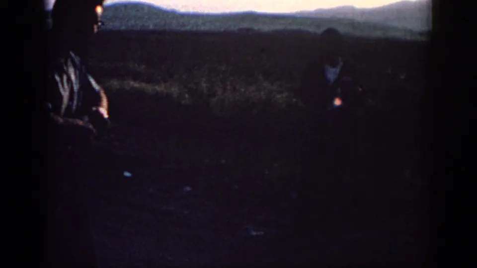 Two adult men appear in silhouette in a rural Rhodes field, with low hills and dim evening light in the background.