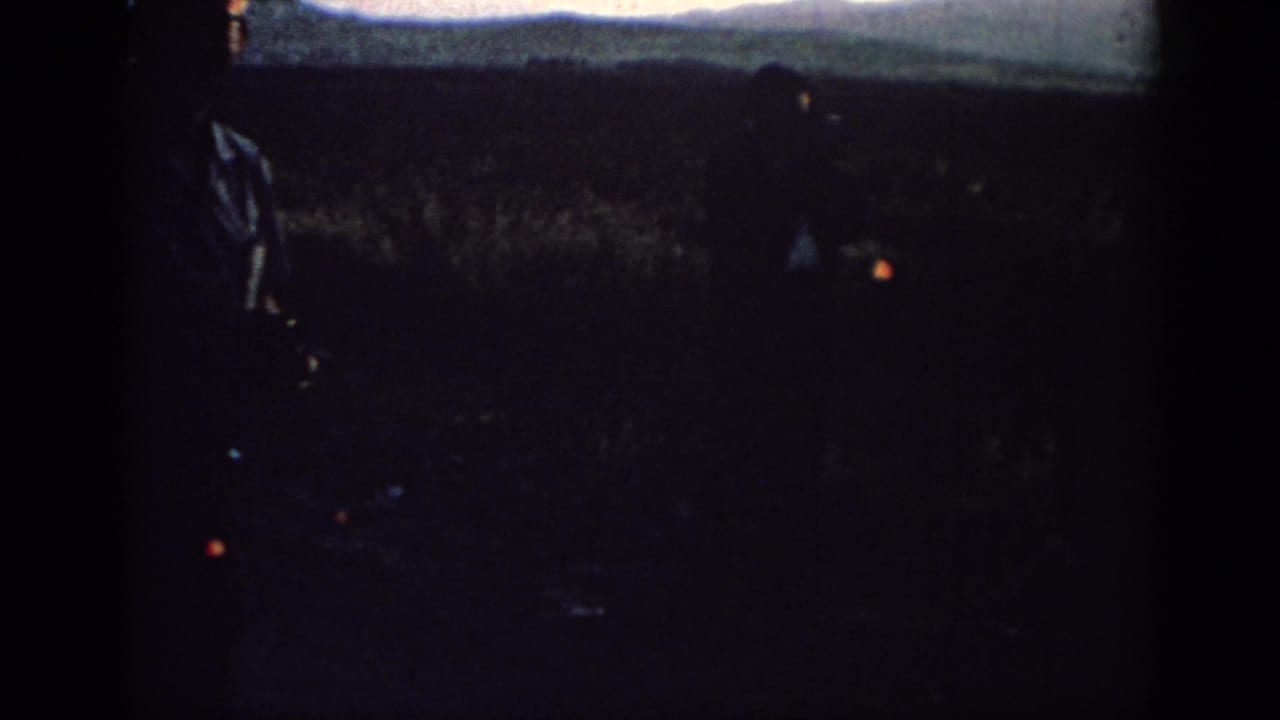 A foreground man and a second distant figure are silhouetted in a Rhodes field at sunset, with the horizon faintly visible.