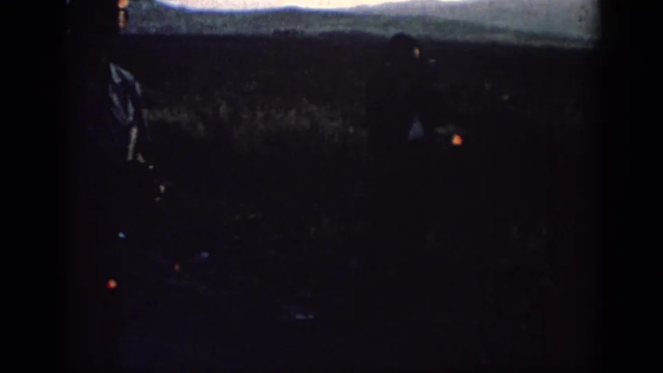 A foreground man and a second distant figure are silhouetted in a Rhodes field at sunset, with the horizon faintly visible.