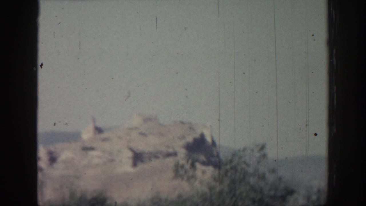 1962 Rhodes home-movie frame with blurred hilltop ruins, foreground scrub, and visible 8mm dust and scratches.