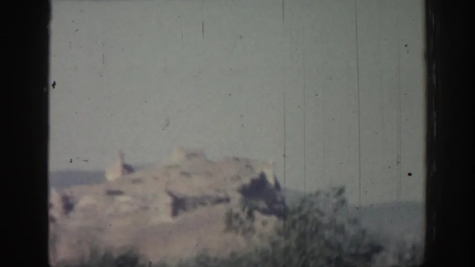 1962 Rhodes home-movie frame with blurred hilltop ruins, foreground scrub, and visible 8mm dust and scratches.
